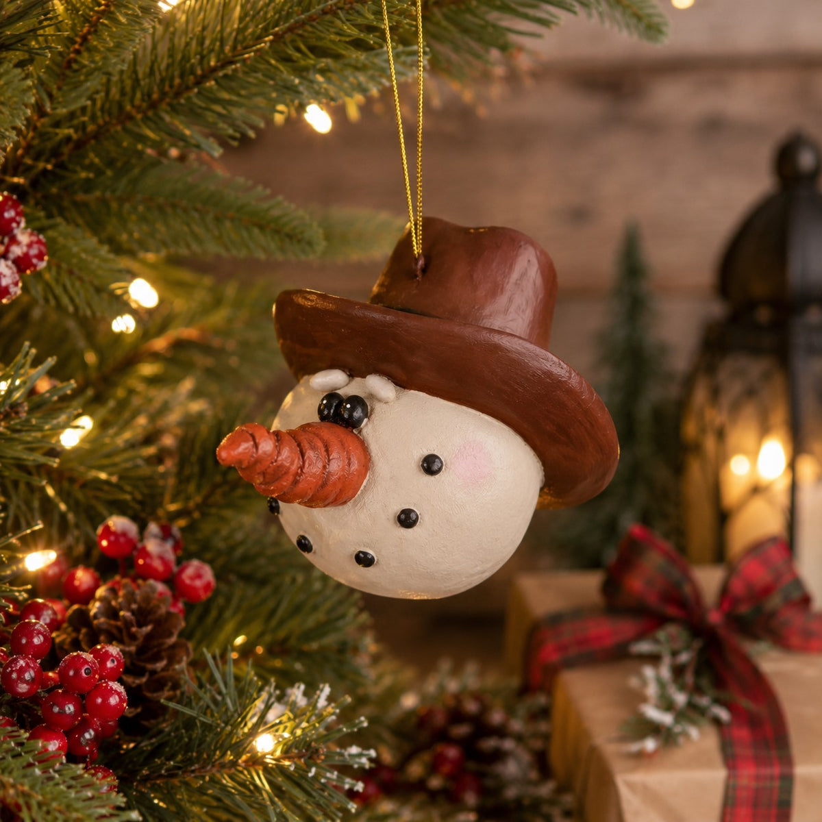 A resin snowman ornament hanging from a gold string on a Christmas tree, wearing a brown cowboy hat and orange carrot nose, with warm holiday lights and wrapped gifts in the background.