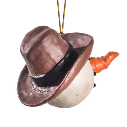 A snowman Christmas ornament wearing a brown cowboy hat with a black band and an orange carrot nose, hanging by a gold string against a white background.