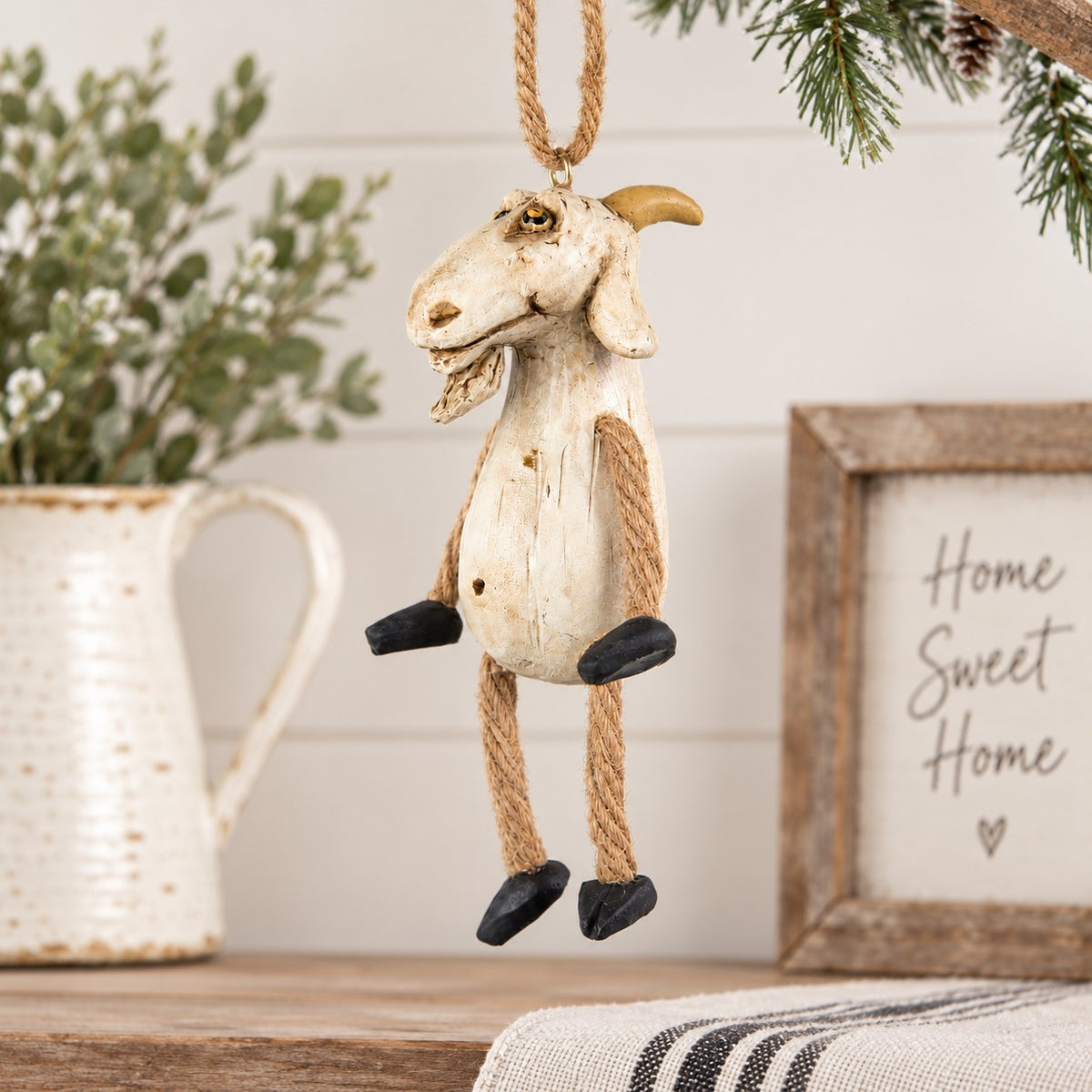 A rustic goat hanging ornament with a distressed cream body, tan rope legs and hanger, and black hooves, displayed on a tabletop with holiday decor in the background.