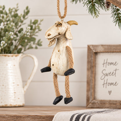 A rustic goat hanging ornament with a distressed cream body, tan rope legs and hanger, and black hooves, displayed on a tabletop with holiday decor in the background.
