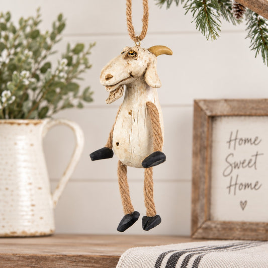 A rustic goat hanging ornament with a distressed cream body, tan rope legs and hanger, and black hooves, displayed on a tabletop with holiday decor in the background.