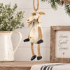 A rustic goat hanging ornament with a distressed cream body, tan rope legs and hanger, and black hooves, displayed on a tabletop with holiday decor in the background.