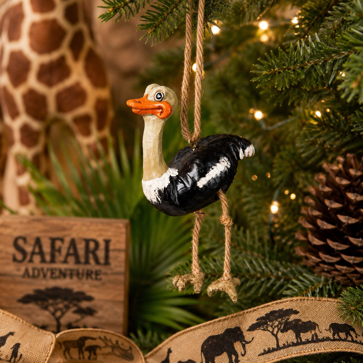 A safari-themed ostrich Christmas ornament hanging from a decorated Christmas tree. The ornament features a black and white body with a beige neck and orange beak and  dangly rope legs,
