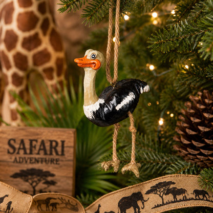 A safari-themed ostrich Christmas ornament hanging from a decorated Christmas tree. The ornament features a black and white body with a beige neck and orange beak and  dangly rope legs,