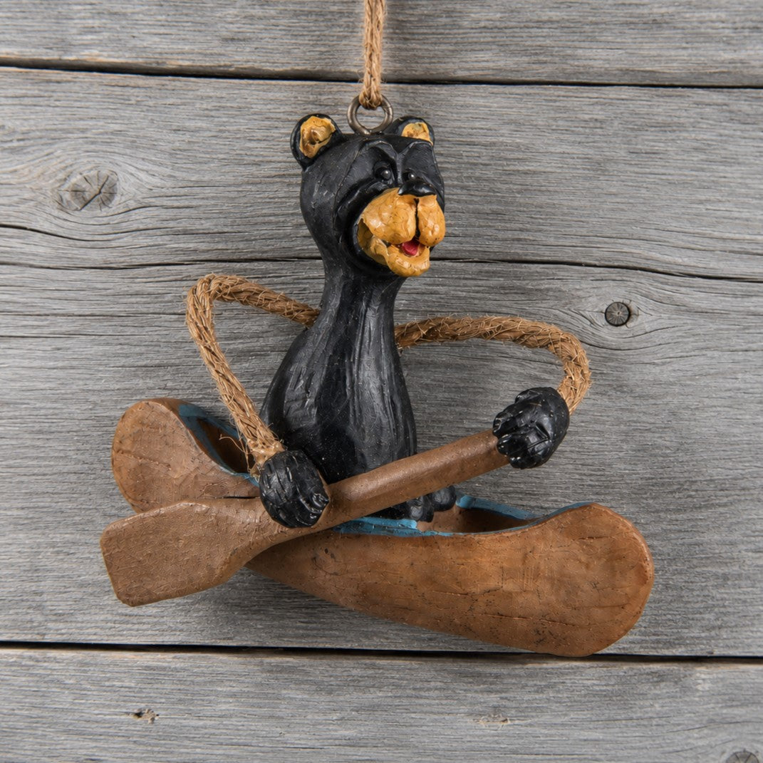 A hand painted black bear ornament sitting in a canoe with a oar crossed over the canoe hanging against a weathered gray wood plank background.