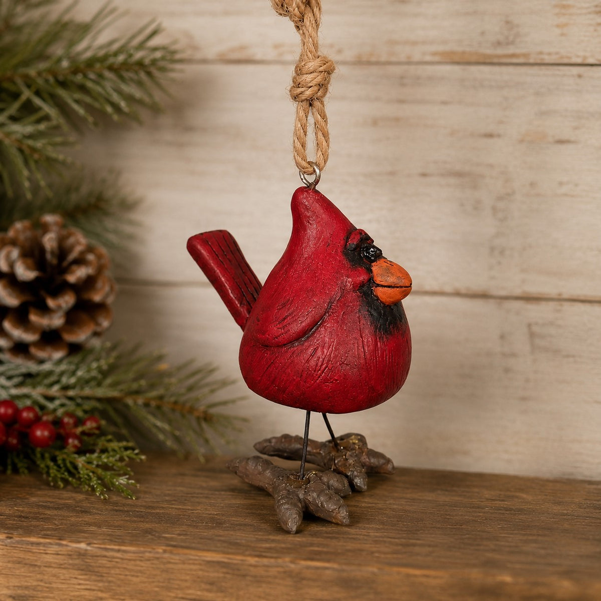 A resin cardinal ornament with wire legs and jute hanger is displayed with evergeen  and pinecones with a rustic wood background.