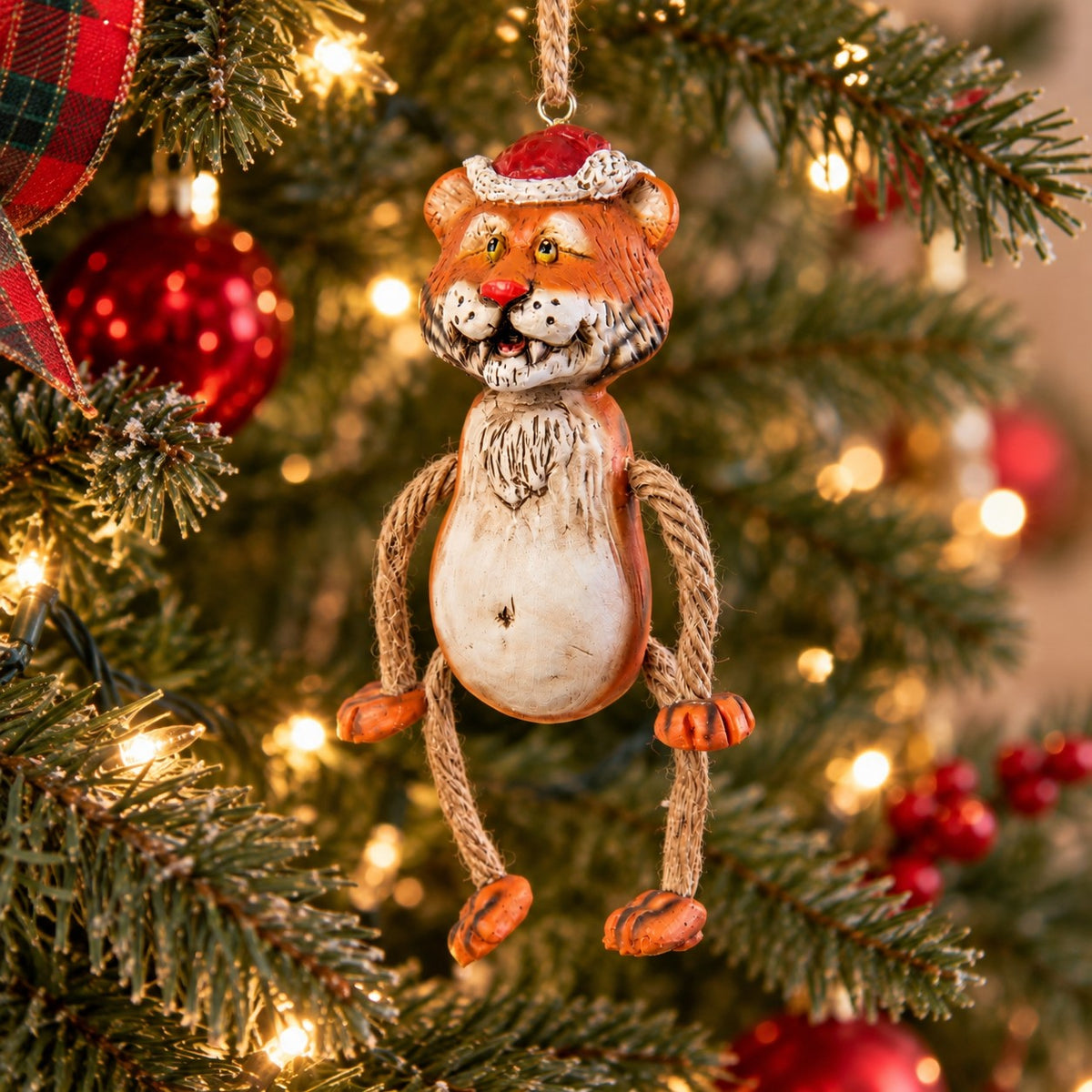Bert Anderson tiger Christmas ornament wearing a Santa hat with dangly rope arms and legs hanging on a lighted Christmas tree.