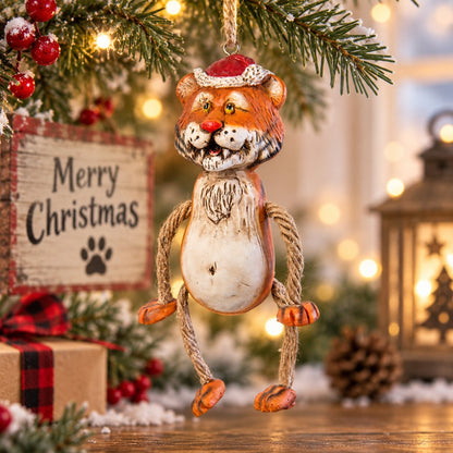 A hanging Tiger Christmas ornament with a Santa hat and jute arms and legs that dangle.  Displayed hanging from a Christmas tree with a festive holiday background.