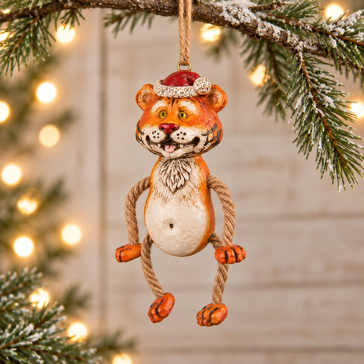 Bert Anderson tiger Christmas ornament wearing a Santa hat with dangly rope legs, displayed with evergreen branches and warm holiday lights.