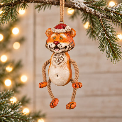 Bert Anderson tiger Christmas ornament wearing a Santa hat with dangly rope legs, displayed with evergreen branches and warm holiday lights.