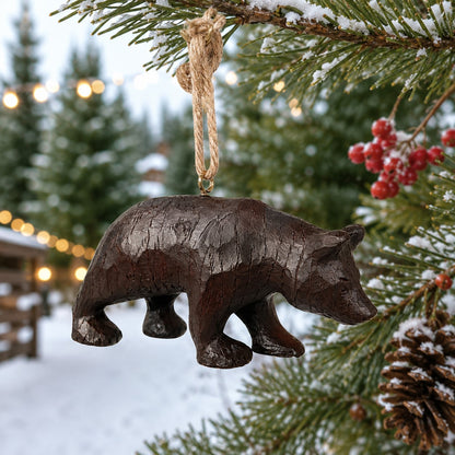 A dark brown bear ornament hanging from a jute rope on a snowy Christmas tree branch, with a blurred winter outdoor background.  
