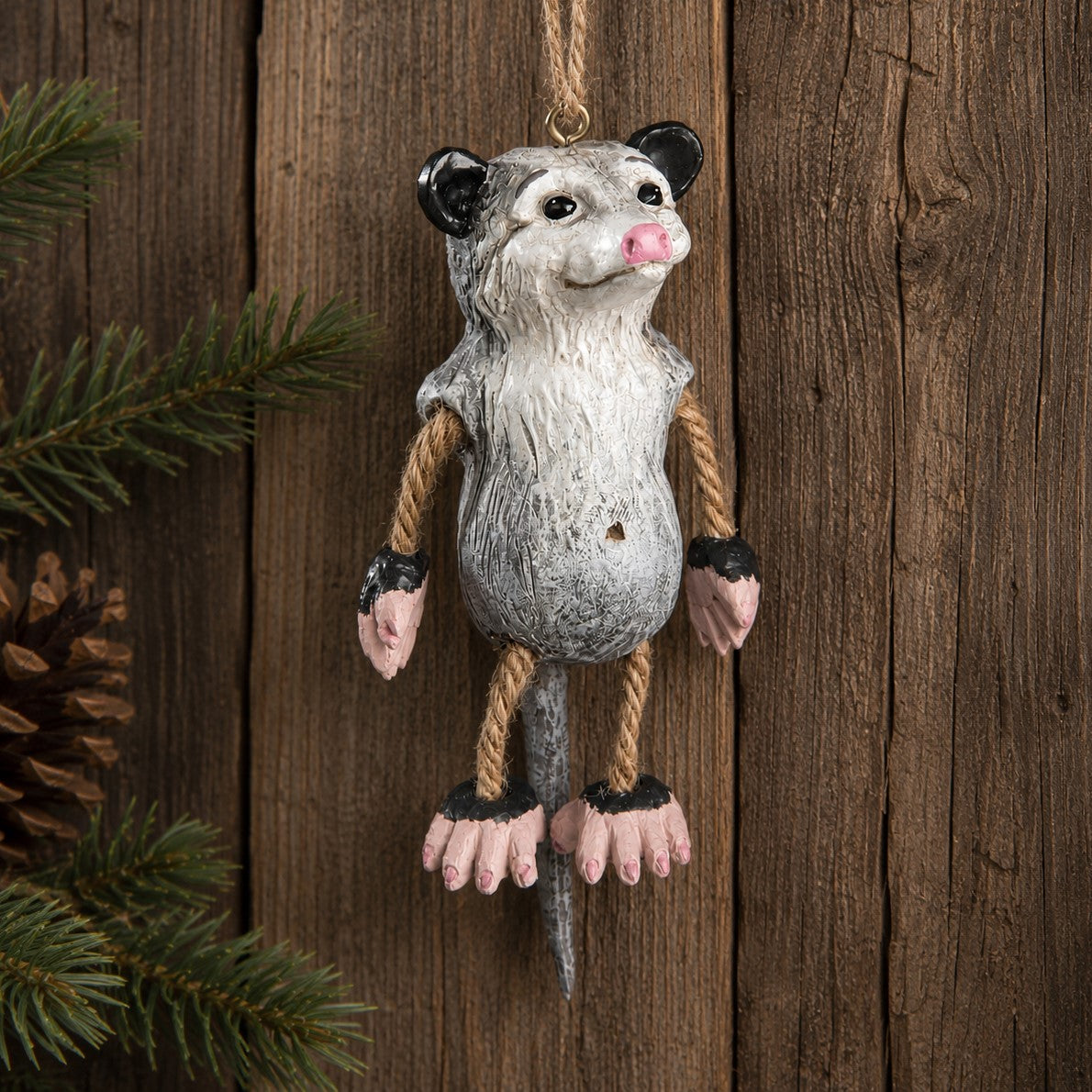 Possum  ornament with  dangly rope arms and legs displayed on a rustic wood wall with evergreen and pinecone.