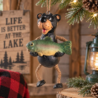 Bert Anderson black bear fishing ornament holding a fish with dangly rope legs, displayed in a rustic Christmas cabin setting.