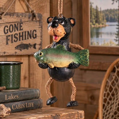 Bert Anderson black bear fishing ornament holding a fish with dangly rope legs, displayed in a rustic lake cabin setting.