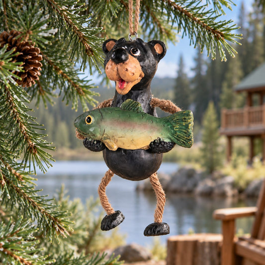 Bert Anderson black bear fishing ornament holding a fish with dangly rope legs, hanging from evergreen branches beside a rustic lakeside cabin.