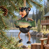 Bert Anderson black bear fishing ornament holding a fish with dangly rope legs, hanging from evergreen branches beside a rustic lakeside cabin.