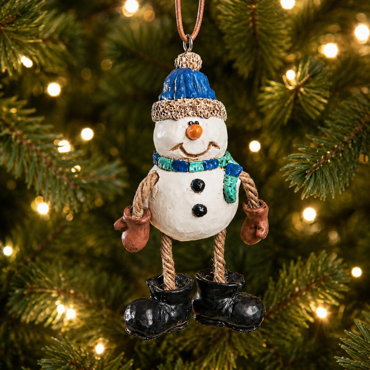 Dangly snowman ornament with blue hat and scarf, rope arms and legs, mittens and oversized black boots hanging on a lit Christmas tree.