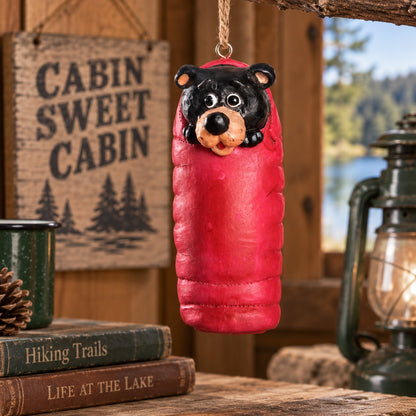 Bert Anderson black bear ornament tucked into a red sleeping bag, displayed in a rustic cabin setting with camping and lake décor.