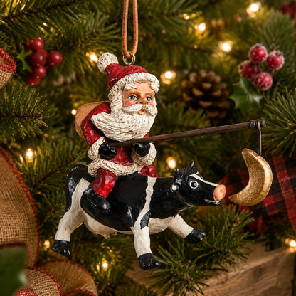 Bert Anderson Santa Riding a Cow ornament with fishing pole and dangling crescent moon, hanging on a Christmas tree with evergreen branches and warm lights.