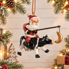 Bert Anderson Santa riding a black-and-white cow Christmas ornament, displayed against a light holiday background with evergreen, red berries and warm lights.