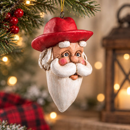 Bert Anderson Cowboy Santa head ornament with red Western hat and white beard hanging in a warm Christmas setting with evergreen and soft holiday lights.
