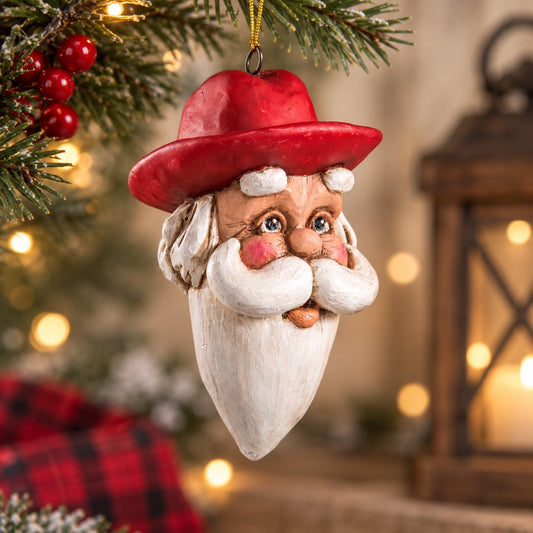 Bert Anderson Cowboy Santa head ornament with red Western hat and white beard hanging in a warm Christmas setting with evergreen and soft holiday lights.