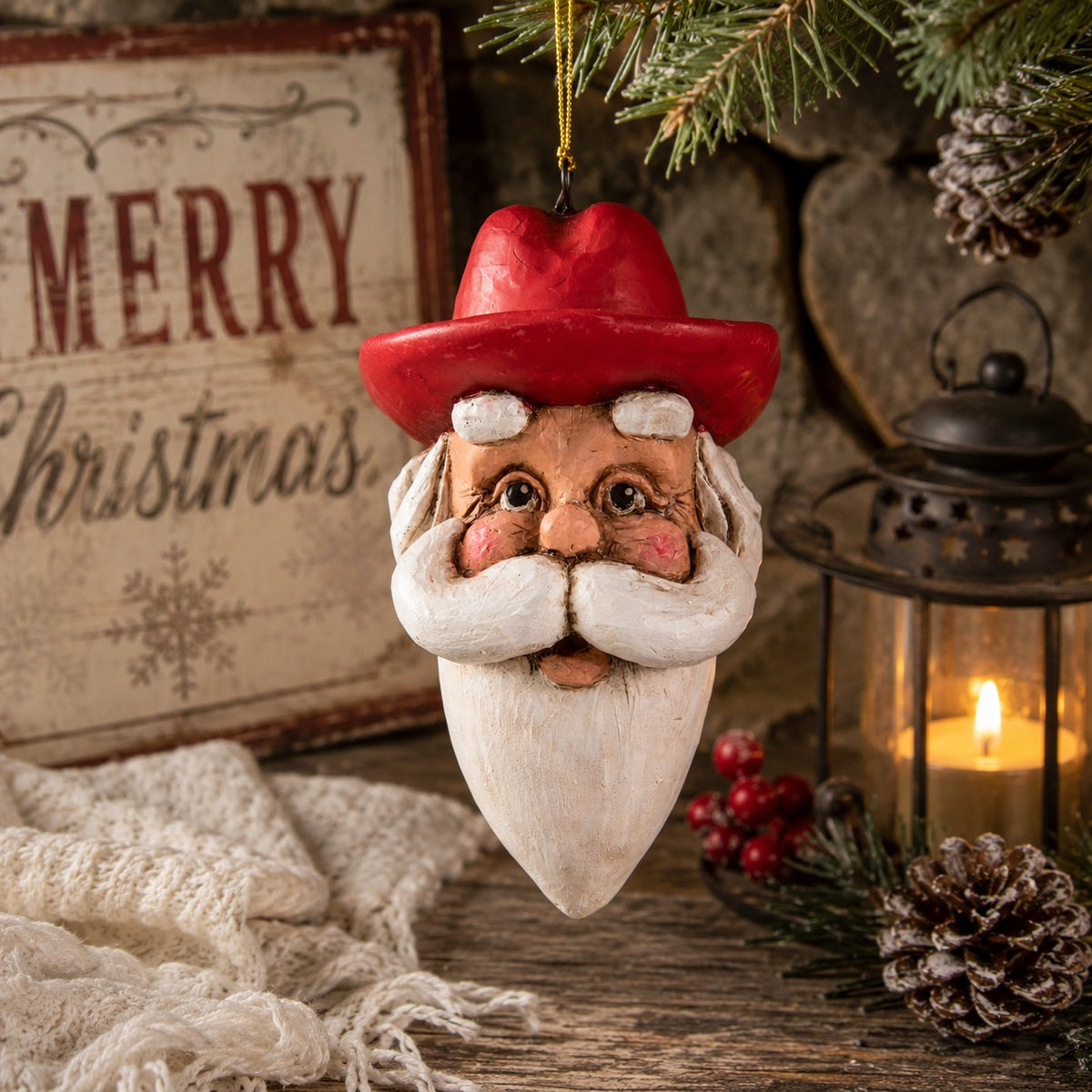 Bert Anderson Cowboy Santa head ornament with red cowboy hat and white beard hanging in a rustic Christmas setting with evergreen and lantern.