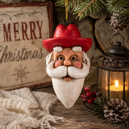 Bert Anderson Cowboy Santa head ornament with red cowboy hat and white beard hanging in a rustic Christmas setting with evergreen and lantern.