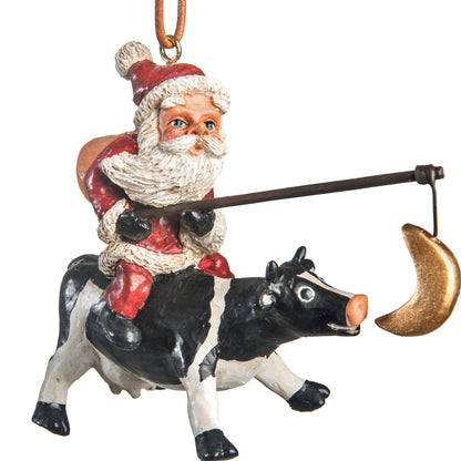 Bert Anderson Santa Riding a Cow ornament with Santa holding a fishing pole and dangling crescent moon on a white background.